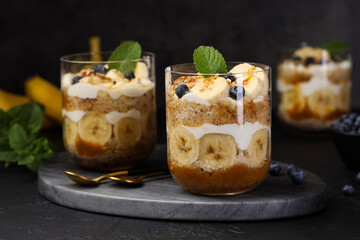 Tasty trifle dessert. Sponge cake, blueberries, banana and whipped cream in glasses on black table