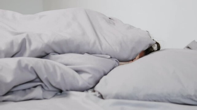 Close-up of a young woman wrapped in gray bedding, lying in bed, unwilling to wake up in a minimalist room.