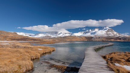 Wooden path leads to tranquil lake with snow-capped mountains under a bright blue sky.