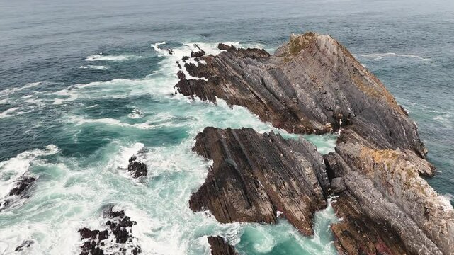 Dramatic view of a large rock formation in the ocean with rough waves crashing against it. Ribat da Atalaia, Portugal