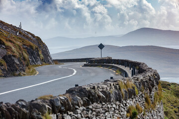 Coastal road winds through rugged Irish landscape, edged by stone walls.  Whitish-gray hills and a...