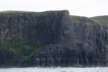 Dramatic, dark cliffs rise sharply from the ocean, layered with strata and small vegetation.  Waves...