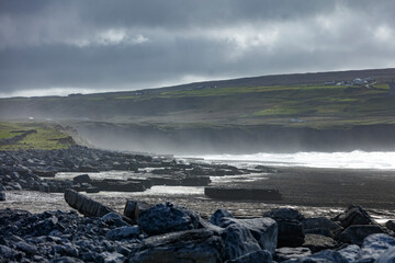 Coastal landscape with rugged, dark gray rocks, a misty shoreline, and a hillside with scattered houses.  Cloudy day.
