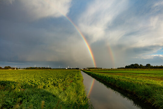 Double rainbow arches over fields and a canal.  Calm, rural scene.