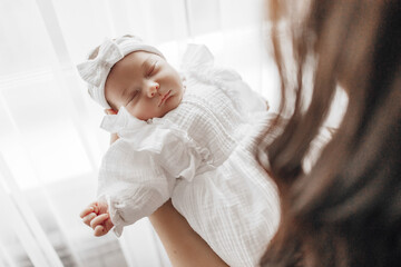 A baby is sleeping in a white dress with a bow on her head. The baby is being held by a woman