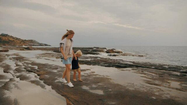 Relaxed family mother and son baby boy walking at stone beach windy weather dramatic sky slow motion