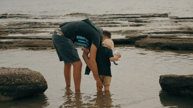 Caring father rolling up pants to son child standing sea water stone beach admiring nature seascape