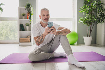 Elderly man in activewear relaxing indoors while using his smartphone on a yoga mat, surrounded by greenery and natural light