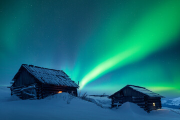 Old wooden cabins in snowy mountains under incredible Northern lights in starry sky. Christmas...