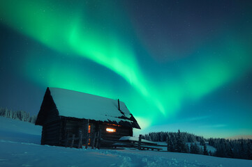 Old wooden cabin in snowy mountains under incredible Northern lights in starry sky. Christmas...