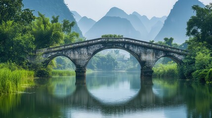 Over the calm river, an old rice bridge, made from weathered stones, connects two sides of a lush, green valley.