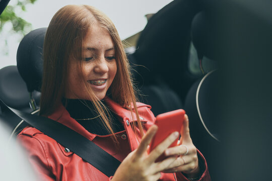 Trendy girl sitting in a convertible car with security belt. Smiling teen using smartphone and chat with friend. Young fashion woman enjoying free time wearing red leather jacket Youth freedom concept - Powered by Adobe