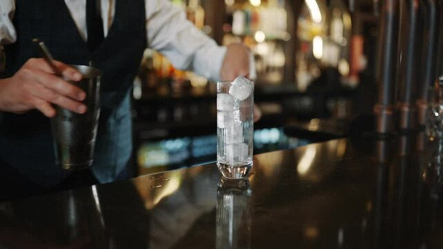 A bartender is making a drink with ice cubes in a glass. The bartender is wearing a black vest and a white shirt