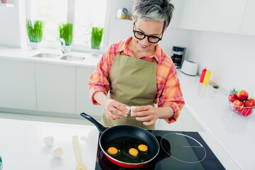 Charming senior woman enjoying cooking in a bright kitchen, cracking eggs into a skillet for...