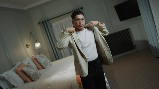 A man in a white shirt and tan jacket is standing in front of a bed. He is adjusting his jacket and he is in a hurry