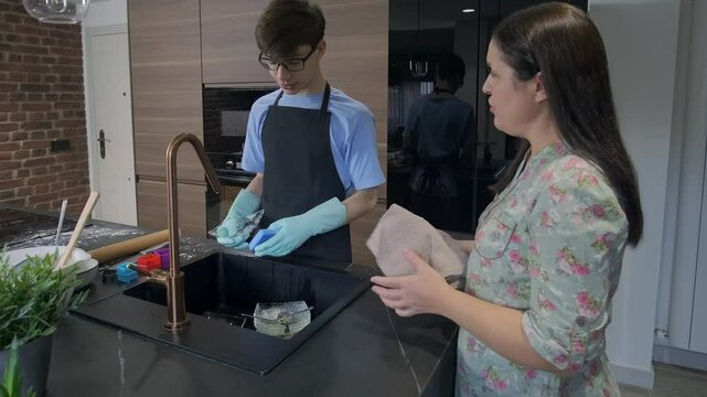Happy family, mother and teenage son wearing gloves washing and drying dishes together in modern kitchen after baking, sharing housework and quality time.