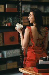 In a warm and inviting setting adorned with colorful gift boxes, a graceful woman in a flowing red dress holds a delicate bowl, exuding charm and poise while enjoying the festive atmosphere
