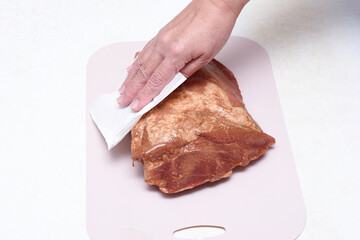A woman's hand removes excess moisture from marinated meat using a paper napkin.