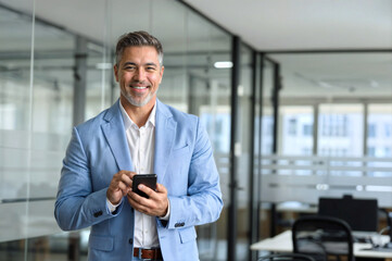 Happy mature business man investor wearing suit standing in office using mobile phone. 50 year old smiling businessman professional holding smartphone looking at camera at work. Portrait, copy space.