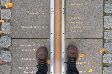 Feet straddling the Prime Meridian at Greenwich, London, with longitude markers engraved on the...