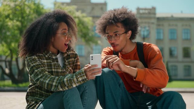 Two multiracial friends students couple African American man guy male with woman girl female together looking mobile phone amazed shock excited good news online achieve win in city outdoors university
