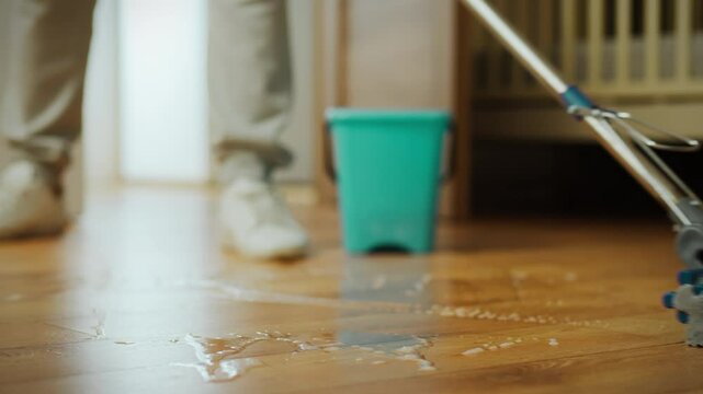 A person is actively cleaning wooden floors using a mop and a bucket. The sunlight filters in through a nearby window, illuminating the tidy living area