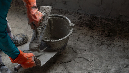 A worker uses a trowel to scoop up cement from a black plastic bucket while plastering a house wall. Concept of working in a construction site