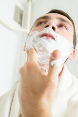Young man applying shaving foam near mirror in bathroom