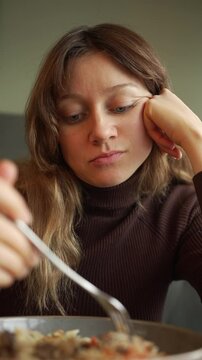 Solitary young woman wearing brown turtleneck sitting at table, languidly pushing lunch around plate with fork, expressing profound disinterest and emotional detachment during midday meal