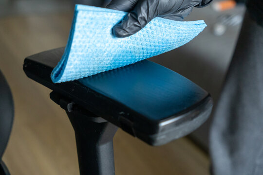 cleaning office chair armrest with blue microfiber cloth, close-up of hand in black gloves during disinfection
