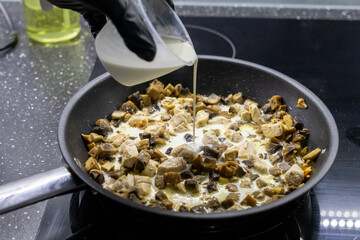 pouring cream into a skillet with chicken, mushrooms, and spices on a stovetop