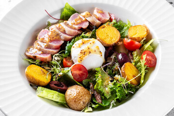 A vibrant salad featuring tender slices of meat, a poached egg, mixed greens, cherry tomatoes, olives, and roasted potatoes, drizzled with a flavorful mustard dressing. Fresh and appetizing.