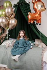 Cheerful little birthday girl 4-5 years in stylish rustic style dress sits on bed decorated with baldachin, pillows and balloons, smiles and looks at camera. Child birthday party celebration. © Andriy Medvediuk