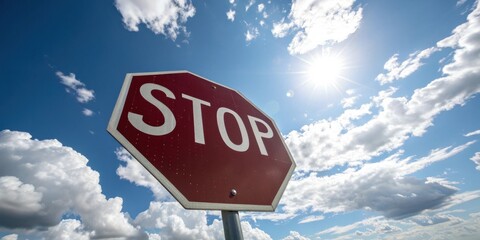 A red stop sign against a bright blue sky with puffy clouds and the sun shining brightly