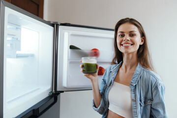 Young woman smiling while holding a green smoothie in front of an open refrigerator filled with fresh vegetables, representing healthy eating and lifestyle choices