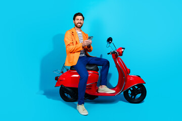 Young man in a bright orange jacket sitting on a red scooter against a turquoise background, smiling confidently