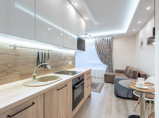 Modern Minimalist Home Kitchen Interior With Wooden Fixtures and Contemporary Appliances. Dining Table and Living Room Area in Background