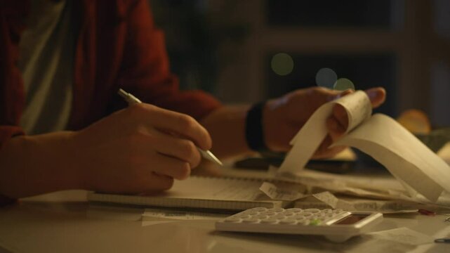 A person works late into the night, surrounded by papers and a calculator, as they meticulously organize and manage their finances while using a pen on a notebook