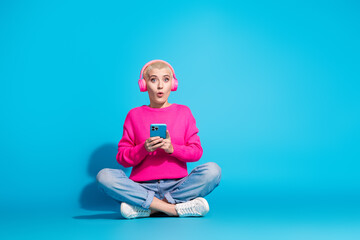 Young woman with a shocked expression seated against a bright blue background, wearing pink headphones