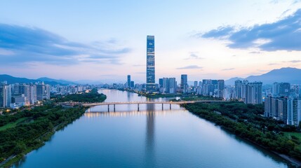 Obraz premium Tall modern skyscraper rising over a river and bridge in a city skyline at dusk with mountains in the background.