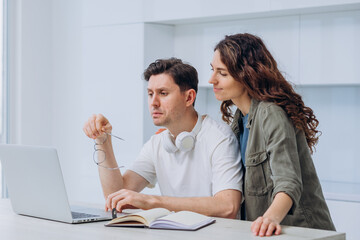 Young man and woman collaborating while working from home, using a laptop and jotting down notes in a notebook, seated at a table in a bright, white kitchen