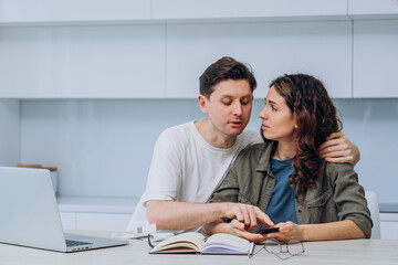Obraz premium Young couple sitting at kitchen table with laptop, notebook, and calculator, managing home finances and calculating expenses together, concerned about their financial situation