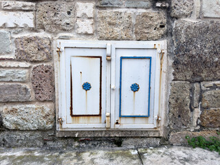 A close-up image of a white-painted iron sliding window, adorned with blue floral patterns, set against an old stone wall. The rusted edges of the window and the weathered masonry highlight the charm 