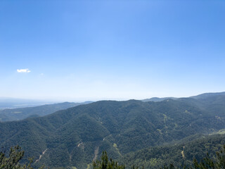 A breathtaking landscape featuring vast, green forest-covered hills under a bright, clear blue sky. A single fluffy white cloud floats in the otherwise spotless sky
