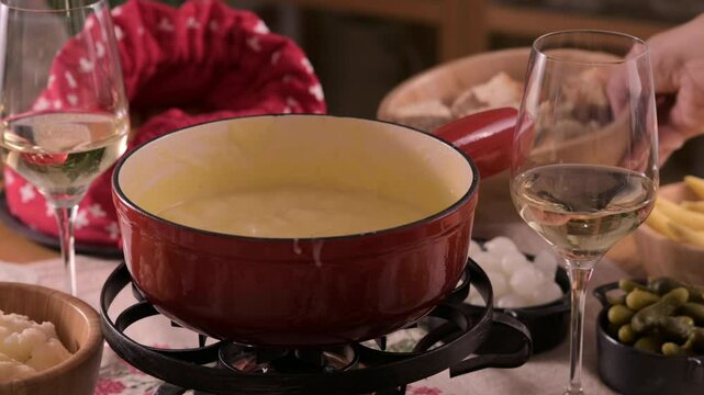 Cozy Swiss fondue setup with melted cheese, bread, pickles, and wine on a rustic table. advertising, social media campaigns, or food-related promotions. High quality 4k footage