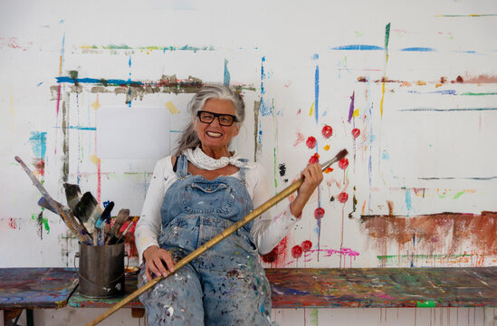 Crazy older mature woman artist, gray hair and glasses sits joyfully at a colorful painted wall, holding a large brush. Her denim overalls reflect a life filled with creativity and passion