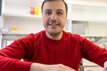 Man wearing a red sweater smiles while sitting at a desk, engaged in a conversation in a well-lit, contemporary lab office space