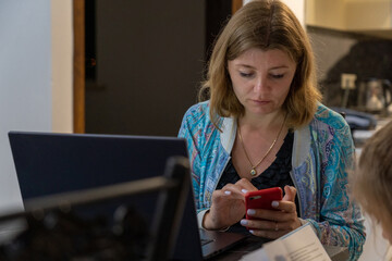 A woman sits in a contemporary kitchen, focused on her smartphone while her laptop is open nearby, indicating multitasking at home