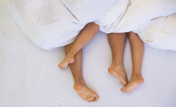 couple lies intertwined under soft white bedding, their legs playfully tangled. The scene conveys intimacy, relaxation, and warmth, suggesting a moment of comfort and connection, copy space