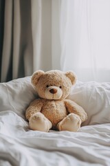 A teddy bear is sitting on a bed. The bear is brown and has black eyes. The bed is white and the room is dimly lit